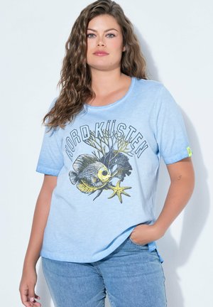 Frau mit welligem braunem Haar, die ein hellblaues T-Shirt trägt, das einen Fisch, Seegras, einen Seestern und den Schriftzug "NORDKÜSTEN" zeigt.