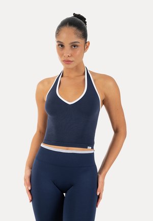 Kobieta z włosami upiętymi w kok, ubrana w granatowy krótki top typu halter z białą lamówką oraz dopasowane legginsy z wysokim stanem na jednolitym tle.