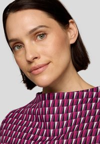 Figurbetontes Oberteil mit einem geometrischen Muster in Rosa, Schwarz und Weiß. Hoher Kragen und strukturierter Stoff. Nahaufnahme des Gesichts des Modells.