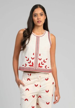 Femme portant un haut sans manches blanc et un pantalon avec des motifs de feuilles rouges brodées, debout avec une main dans la poche devant un fond uni.