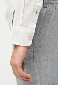 Άνδρας με χαλαρό χέρι στο πλάι που φοράει λευκό πουκάμισο με μανσέτα κουμπωμένη και γκρι υφασμάτινο παντελόνι.