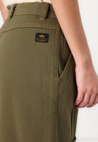 Jupe vert olive en denim de coton, avec une poche latérale et une étiquette noire avec un texte jaune à l'arrière. Coutures visibles accentuées.