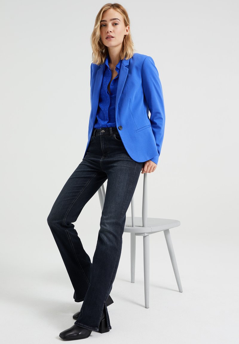 Blauer Blazer, hellblaues Hemd und dunkle Jeans. Der Blazer hat einen Knopf und einen strukturierten Schnitt. Die Person steht neben einem grauen Stuhl.