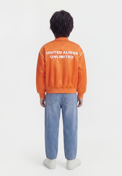 Άτομο με πορτοκαλί μπουφάν με την επιγραφή "UNITED ALIENS UNLIMITED", ανοιχτό μπλε τζιν και λευκά παπούτσια, γυρισμένο πλάτη, σε απλό φόντο.