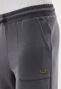 Σκούρο γκρι φόρμα με ελαστική μέση, πλαϊνή τσέπη και μικρό χρυσό λογότυπο EA7 Emporio Armani κοντά στην τσέπη.