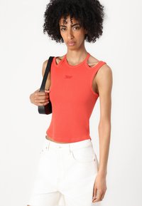 Rot gestreifter Tank-Top mit einzigartigen Schulterausschnitten, ausgestattet mit einem Logo-Detail und kombiniert mit weißen Denim-Shorts und einem schwarzen Schulterriemen.