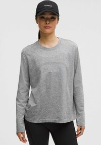 Maglietta grigia a maniche lunghe realizzata in tessuto morbido. Presenta un colletto tondo e una vestibilità comoda. Abbinata a un cappellino nero e leggings scuri.