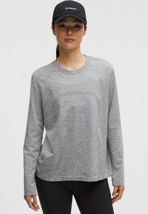 Graues Langarmshirt aus weichem Stoff. Mit Rundhalsausschnitt und lockerer Passform. Kombiniert mit schwarzer Kappe und dunklen Leggings.