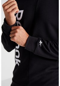 Sudadera negra con un audaz logo blanco de "Reebok" en el lateral y un pequeño emblema blanco en el puño. El material parece suave y texturizado.