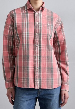 Person wearing a pink and gray plaid long-sleeve button-up shirt with a collar and blue jeans, standing against a light background.
