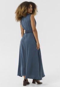 Robe longue sans manches en tissu bleu foncé doux. Présente une taille ajustée liée par une ceinture. Le mannequin porte des chaussures à talons épais marron.