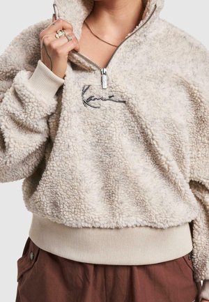 Osoba nosí béžový sherpa fleece pulovr s polovičním zipem a vyšívaným logem, v kombinaci s hnědými kalhotami s vysokým pasem.