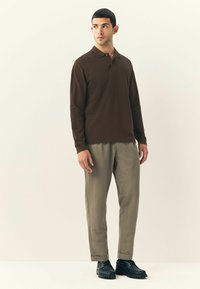 Brązowa koszula polo z długim rękawem, khaki spodnie i czarne buty. Polo ma kołnierzyk z trzema guzikami; spodnie mają luźny krój i mankiety.