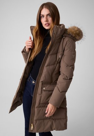 Mujer con abrigo de invierno acolchado marrón con capucha forrada de piel, top negro y vaqueros azul oscuro, de pie frente a un fondo liso.