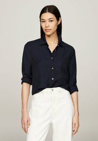 Camicia blu navy con colletto, maniche arrotolate, bottoni dorati e una texture liscia, abbinata a pantaloni bianchi a vita alta.