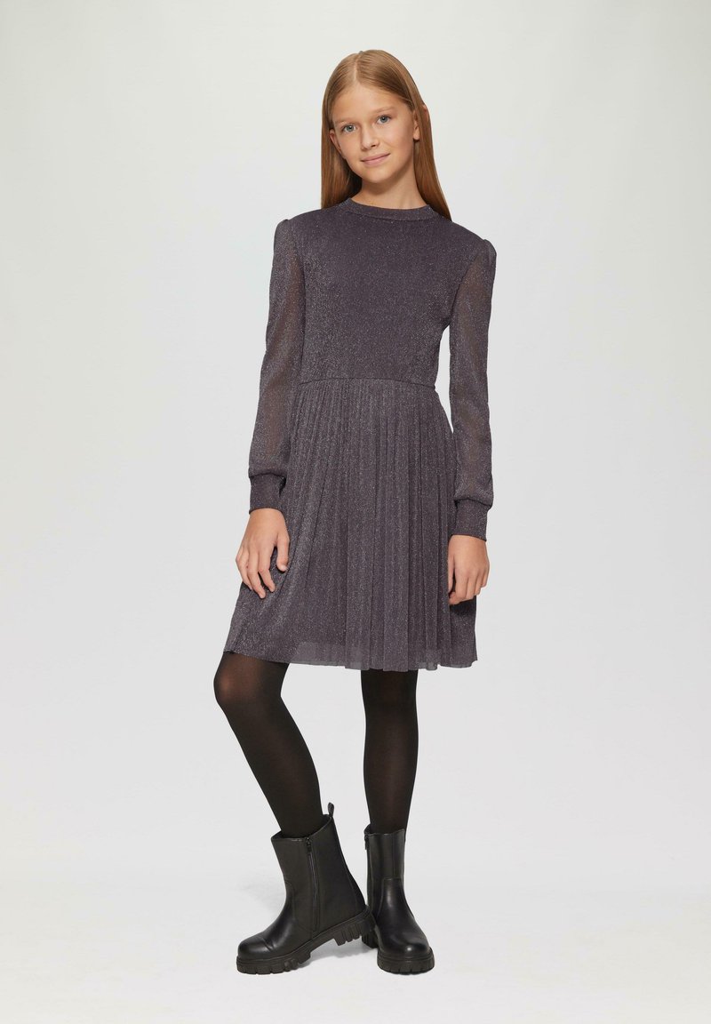 Graues Glitzerkleid mit langen transparenten Ärmeln, gefaltetem Rock, rundem Ausschnitt, kombiniert mit schwarzen Stiefeletten und schwarzen Strumpfhosen.