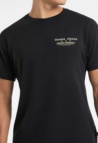 Czarny t-shirt z okrągłym dekoltem i krótkimi rękawami, z małym białym i żółtym logo "Guess Jeans Denim Company" na lewej piersi.
