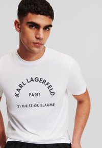 Balta medvilninė marškinėlių su apvalia iškirpte, su juodu spaudiniu, kuriame yra "Karl Lagerfeld" ir adresas.