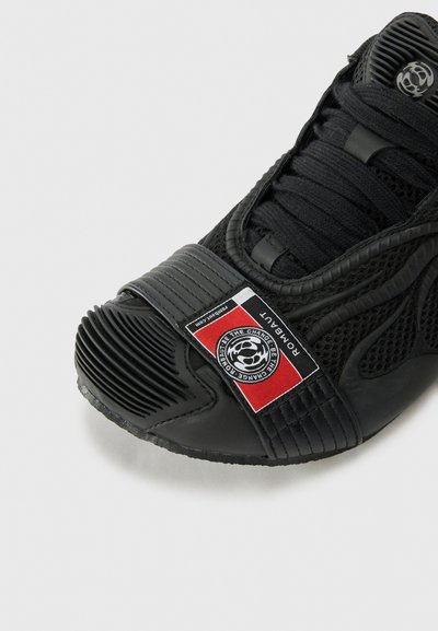 Chaussure de sport noire avec une tige en maille, un capuchon en caoutchouc texturé à l'avant, et une sangle proéminente arborant un badge logo en noir, rouge et blanc.