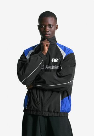 Ung person i en sort og blå zip-up jakke med hvide accenter, som trækker kraven op, står foran en ensfarvet hvid baggrund.