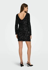 Vestido de lentejuelas negro con mangas largas y un escote en forma de V pronunciado en la espalda. La silueta ajustada resalta una superficie texturizada, complementada con sandalias de tacón alto.