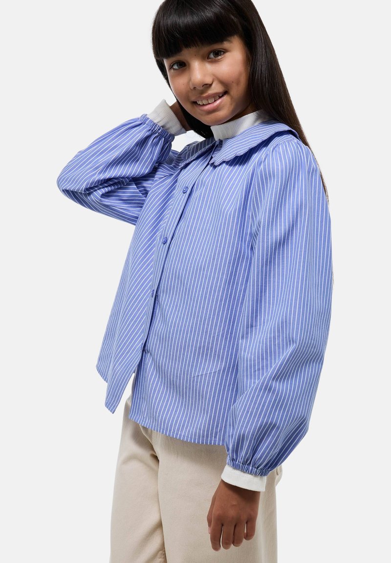 Chemise à rayures bleues et blanches avec un col froncé et des manches amples, associée à un pantalon de couleur claire.