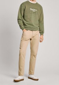 Hombre que lleva un sudadera verde oliva de Pepe Jeans, pantalones beiges y zapatillas blancas, de pie con una mano en el bolsillo.