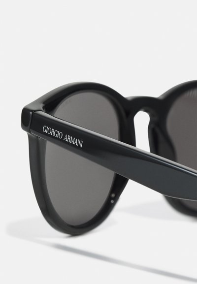 Occhiali da sole tondi neri con lenti grigie, caratterizzati da una finitura lucida, una montatura spessa e il nome "Giorgio Armani" in argento sulla gamba.
