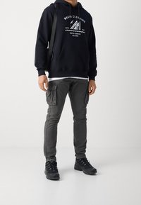 Granatowa bluza z kapturem z białym logotypem, szare spodnie cargo z bocznymi kieszeniami oraz czarne buty sportowe. Prezentacja casualowego stroju.