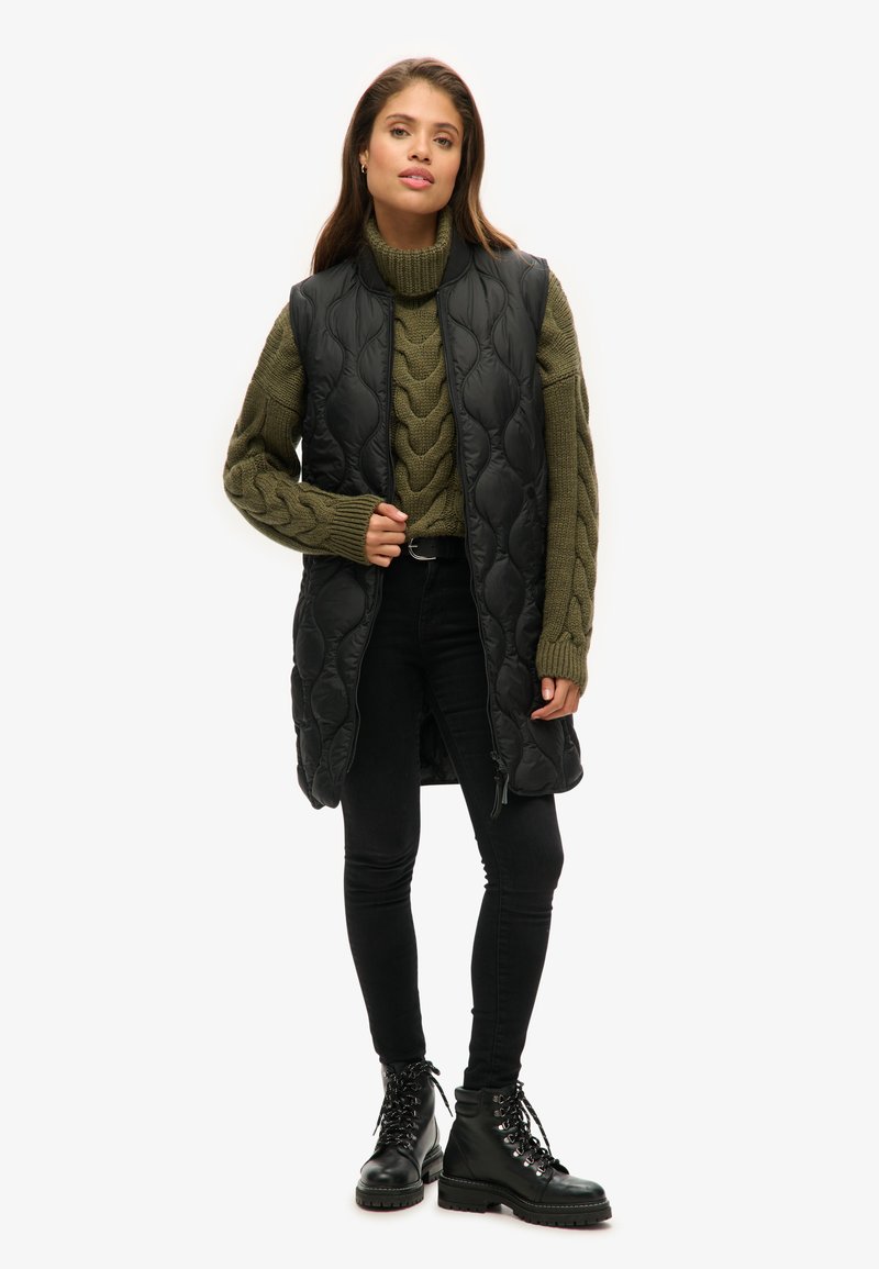 Schwarze gesteppte lange Weste über einem olivgrünen Strickpullover, kombiniert mit schwarzen Skinny-Jeans und schwarzen Schnürstiefeln.