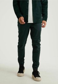 Mörkgröna, slim-fit byxor med en slät yta och diskret lyster, kombinerat med svarta sneakers med en ljusfärgad sula.