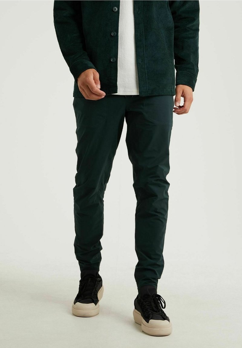 Mörkgröna, slim-fit byxor med en slät yta och diskret lyster, kombinerat med svarta sneakers med en ljusfärgad sula.