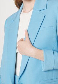 Blazer azzurro chiaro con spalle strutturate, rever a punta e design pulito. Indossato sopra un top cremisi testurizzato, che mostra una singola tasca verticale.