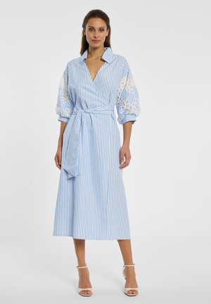 Vrouw staand, gekleed in een midi-jurk met blauw-witte verticale strepen, met pofmouwen van kant en een bijpassende stoffen riem, beige sandalen met hoge hakken.