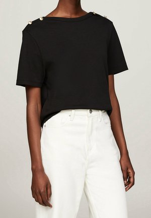 Schwarzes T-Shirt mit kurzen Ärmeln und weitem Rundhalsausschnitt, mit goldenen Hardware-Details an den Schultern. Kombiniert mit weißen, hochgeschnittenen Jeans.