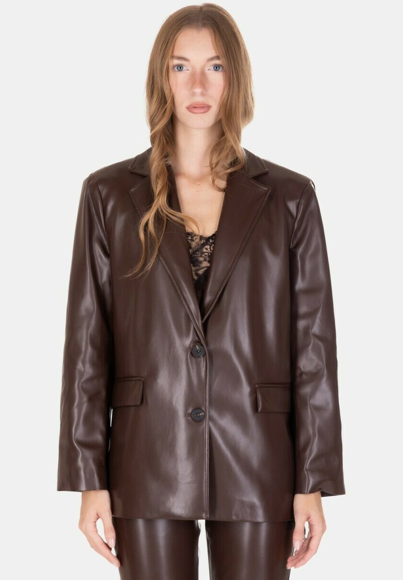 Donna che indossa un blazer in pelle marrone scuro e pantaloni abbinati, con capelli mossi e sciolti e una top in pizzo nero sotto, che si trova davanti a uno sfondo bianco.