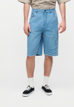 Mand iført lyseblå denim shorts til knæet med frynsede geometriske mønstre, hvide sokker, sorte sneakers og en ternet skjorte.