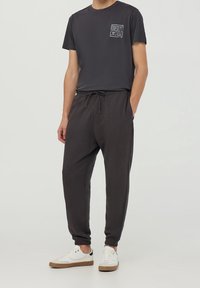 Muž v černém tričku One Piece, tmavých jogger kalhotách s šňůrkou a bílých teniskách s gumovou podrážkou, stojící s jednou rukou v kapse.