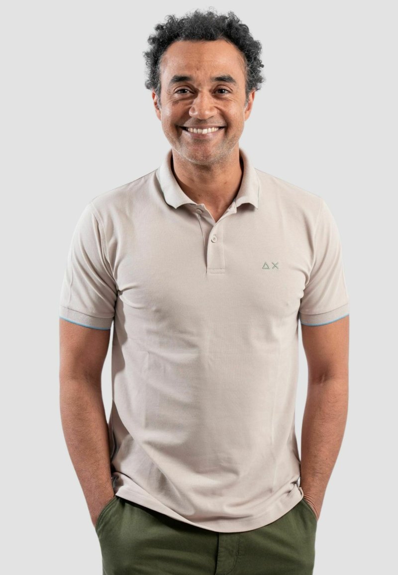Polo shirt beige chiaro con maniche corte, caratterizzato da un piccolo logo verde sul petto e dettagli blu ai bordi delle maniche.