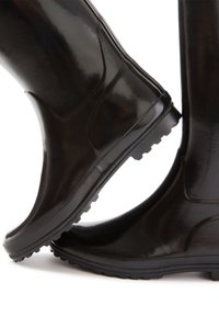 Paar schwarze Gummistiefel mit strukturierten Sohlen, ein Stiefel leicht angehoben und auf dem anderen ruhend vor weißem Hintergrund.