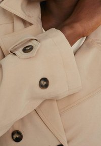 Beżowa kurtka z tkaniny o zdefiniowanym kroju. Posiada duże ciemne guziki oraz przeszycia na mankietach i kołnierzu.