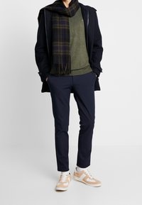 Mies, joka on pukeutunut tummaan takkiin, vihreään villapaitaan, tummansinihin housuihin, ruudulliseen huiviin, valkoisiin sukkiin ja beigeihin tennareihin, seisoo vaaleaa taustaa vasten.