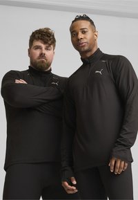 Schwarze Sportshirts mit Reißverschlusskragen, langen Ärmeln und silbernen Logo-Akzenten, kombiniert mit schwarzen Leggings. Glatter, elastischer Stoff.