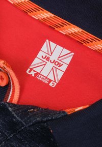 Marineblauwe stof met rode en oranje accenten; heeft een geweven kraag, knoopsluiting en een label met een ontwerp van de Britse vlag.