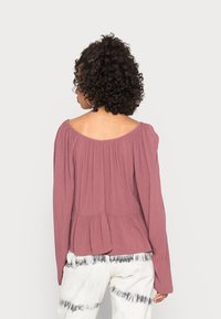 Langærmet off-shoulder bluse i støvet rosa, med plisseret design og peplum-facon, parret med lyse tie-dye bukser.