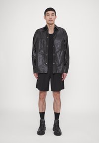Camisa chaqueta de cuero negro con botones a presión, combinada con una camiseta sin mangas negra y pantalones cortos. Botas y calcetas negras completan el atuendo.