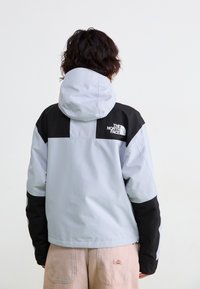 Asmuo su garbanotais plaukais, dėvintis šviesiai pilką ir juodą "The North Face" džemperį bei smėlio spalvos kelnes, atsisukęs nuo objektyvo priešais vientisą foną.