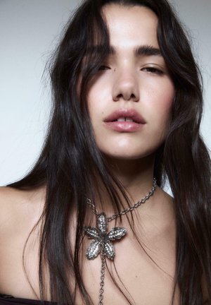 Sömlös silverblomsterhalsband med intrikat design, kedjedetalj och en central blompendant. Slät hud och mörkt hår ramar in smycket.