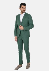 Completo verde strutturato con una vestibilità su misura, caratterizzato da un blazer a un bottone e pantaloni slim, abbinato a una camicia bianca e mocassini grigi.
