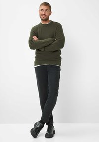 Maglione lavorato a maglia verde oliva con collo rotondo, indossato sopra una camicia bianca. Pantaloni neri attillati e scarpe nere completano l'outfit.